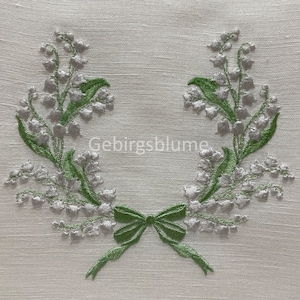 Puede incluir: Una tela de lino blanca con una corona bordada de flores de lirio de los valles en plata y verde.