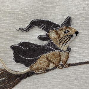 May include: Embroidered design of a brown mouse wearing a black witch hat and riding a brown broomstick.
