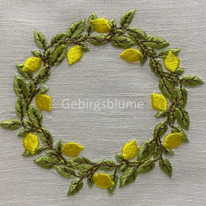 以下が含まれることがあります： 白い布に刺繍されたレモンのリース。黄色いレモンと緑の葉が特徴で、中央には「Gebirgsblume」の文字があります。刺繍は細かく、色は鮮やかです。