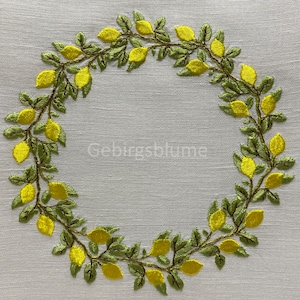 以下が含まれることがあります： 白いリネン製のテーブルクロスに、緑の葉と黄色のレモンの刺繍されたリースが施されています。リースはテーブルクロスの中央に配置され、リースの中央には「Gebirgsblume」という文字が印刷されています。