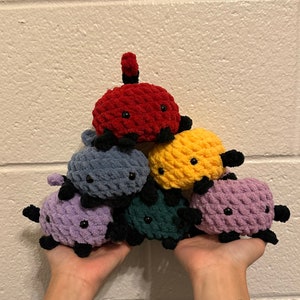 Puede incluir: Una pila de seis juguetes de peluche de crochet en varios colores, incluyendo rojo, amarillo, azul, morado y verde. Los juguetes tienen forma de animales pequeños con ojos negros y están dispuestos en forma de pirámide.