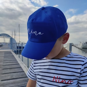 Peut inclure: Une casquette de baseball bleue avec le nom "Nino" brodé en blanc sur le devant. La casquette est portée par un enfant portant un t-shirt rayé avec le nom "Nino" brodé en rouge sur le devant.