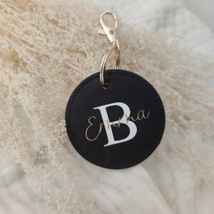 Peut inclure: Porte-clés rond noir avec une lettre "B" blanche et le nom "Emma" écrit en lettres dorées.