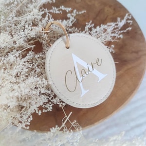 Peut inclure: Un porte-clés rond beige avec un anneau doré. Le porte-clés porte le nom "Claire" en lettres dorées et la lettre "A" blanche. Le porte-clés est posé sur une surface en bois avec des fleurs séchées.