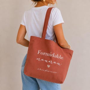 May include: A terracotta-colored tote bag with the words "Formidable nounou" and "et bien plus encore" printed in white. A small heart graphic is below the word "nounou". The bag has long straps for carrying over the shoulder.
