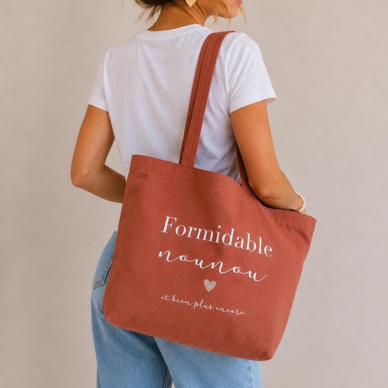 May include: A terracotta-colored tote bag with the words "Formidable nounou" and "et bien plus encore" printed in white. A small heart graphic is below the word "nounou". The bag has long straps for carrying over the shoulder.