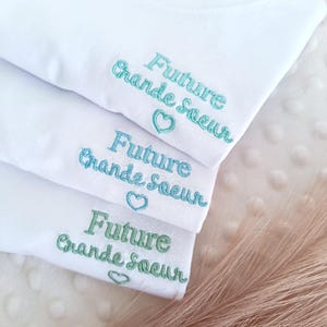 Peut inclure: Trois t-shirts blancs superposés, chacun brodé des mots "Future Grande Soeur" en vert et bleu, avec un symbole de cœur. Les t-shirts sont pliés. Le texte est en français.