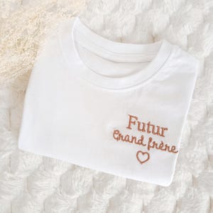 Peut inclure: Un t-shirt blanc à manches courtes avec une broderie marron sur le côté droit. Le texte "Futur grand frère" est écrit en lettres cursives, surmontant le contour d'un petit cœur brodé.