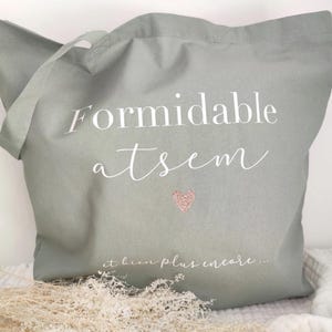 May include: Sage green tote bag with white script text that reads "Formidable atsem" and a small pink glitter heart. The phrase "et bien plus encore..." is written below the heart. The bag has a handle for carrying.