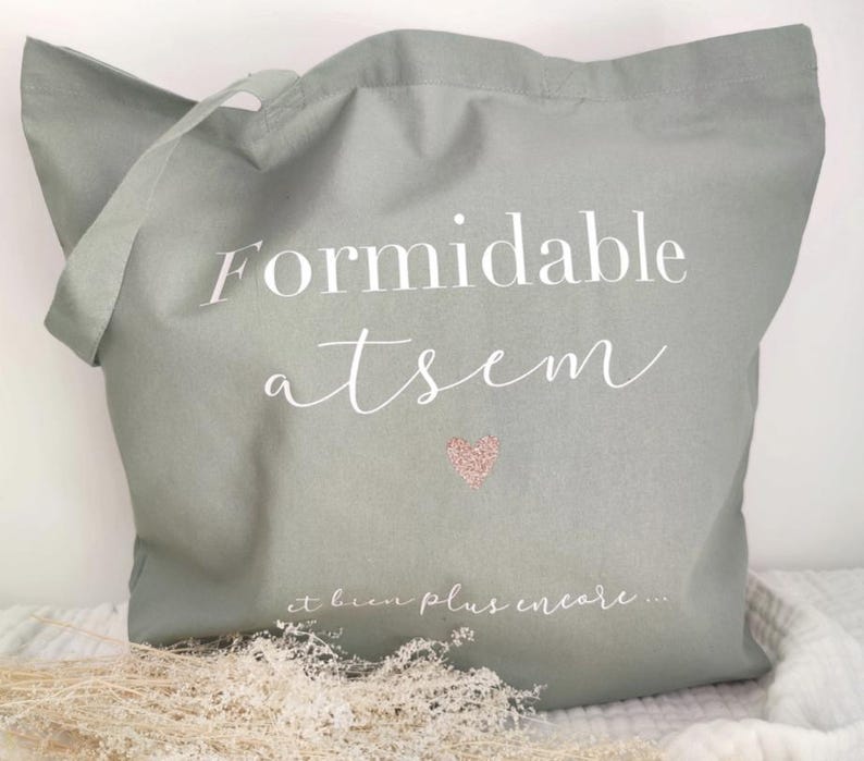 May include: Sage green tote bag with white script text that reads "Formidable atsem" and a small pink glitter heart. The phrase "et bien plus encore..." is written below the heart. The bag has a handle for carrying.