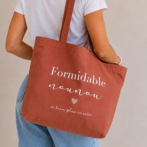 May include: A rust-colored tote bag with the words "Formidable nounou" and "et bien plus encore..." in white script. A small glitter heart is below the text. The bag has long straps.