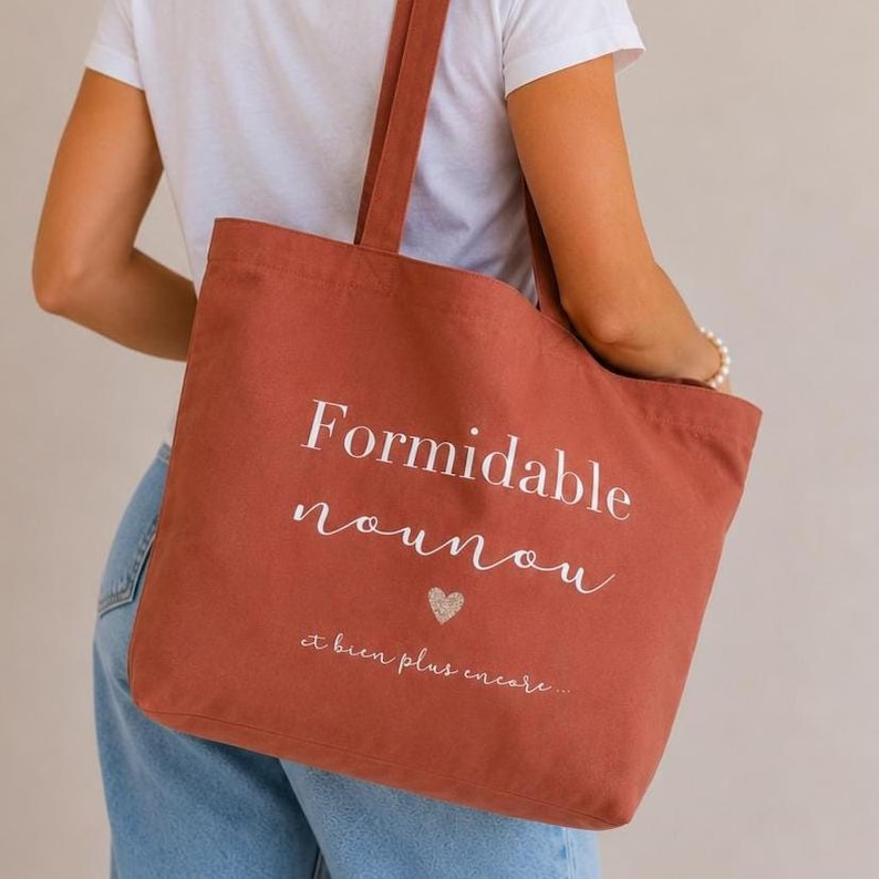 May include: A rust-colored tote bag with the words "Formidable nounou" and "et bien plus encore..." in white script. A small glitter heart is below the text. The bag has long straps.