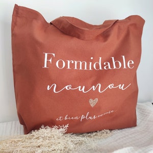 May include: A terracotta-colored tote bag with white text that reads "Formidable nounou" and "et bien plus encore..." A small glittery heart is below the text. The bag has two handles and is made of a sturdy fabric.