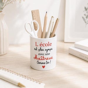 Peut inclure: Un pot à crayons en céramique blanche avec le texte "L'école est plus sympa avec une Maîtresse Comme toi!". Il contient une règle en bois, des ciseaux, des crayons et des stylos. Un petit cœur rouge est en bas.