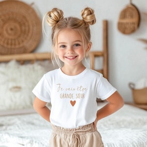 Peut inclure: Une petite fille porte un t-shirt blanc avec l'inscription "Je vais être GRANDE SOEUR" en marron et un petit cœur marron. La fille a les cheveux blonds coiffés en deux chignons et sourit.