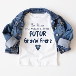 Peut inclure: Un t-shirt blanc avec l'inscription en bleu marine : "Les bêtises c'est mieux à deux ! FUTUR Grand Frère 2026", avec un cœur bleu marine. Une veste en jean est posée sur le t-shirt.