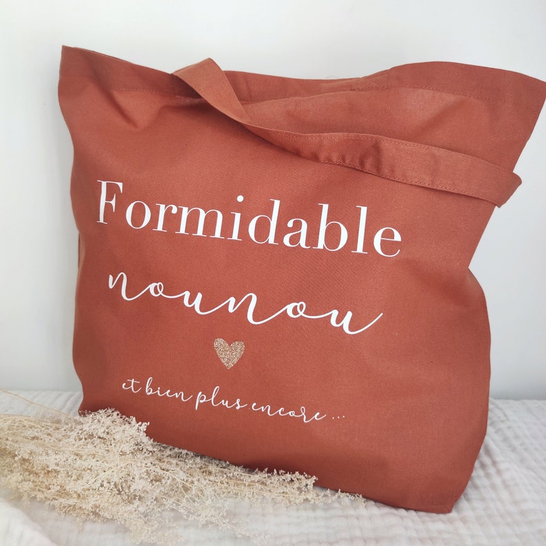 May include: A terracotta-colored tote bag with white text that reads "Formidable nounou" and "et bien plus encore..." with a glitter heart. The bag has a handle and is made of a sturdy fabric.