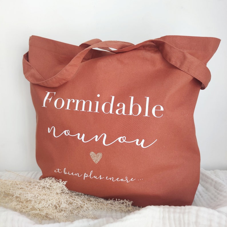 May include: A terracotta-colored tote bag with white text that reads "Formidable nounou" and "et bien plus encore..." with a glitter heart. The bag has two handles and is made of a soft fabric.