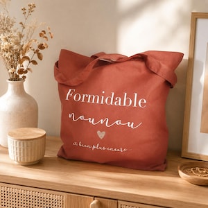 May include: A terracotta-colored tote bag with the words "Formidable nounou" in white script, a small heart, and the phrase "et bien plus encore..." The bag has two handles and is sitting on a wooden surface.