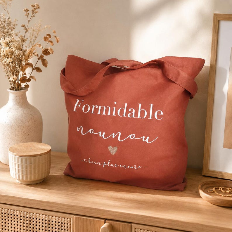 May include: A terracotta-colored tote bag with the words "Formidable nounou" in white script, a small heart, and the phrase "et bien plus encore..." The bag has two handles and is sitting on a wooden surface.