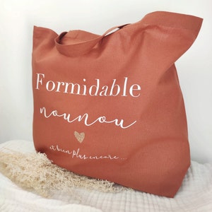 May include: A terracotta-colored tote bag with white script that reads "Formidable nounou" and "et bien plus encore..." with a gold glitter heart. The bag has a handle and is made of a sturdy fabric.