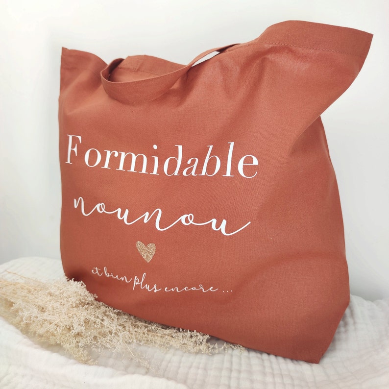 May include: A terracotta-colored tote bag with white script that reads "Formidable nounou" and "et bien plus encore..." with a gold glitter heart. The bag has a handle and is made of a sturdy fabric.