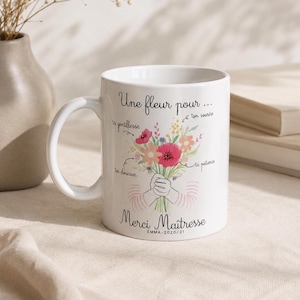 Peut inclure: Mug en céramique blanche avec une anse, orné d'un bouquet de fleurs aux couleurs vives. Le texte comprend "Une fleur pour..." et "Merci Maîtresse" avec un nom et une date. Idéal pour un cadeau.