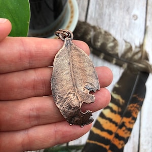 May include: A copper leaf pendant with a detailed, textured surface. The leaf has a small hole in the center and a loop at the top for attaching a chain.