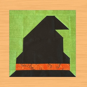May include: A square quilt block featuring a black witch's hat with an orange band decorated with spiderwebs. The hat is set against a green background, creating a Halloween theme. The block is on a light brown surface.