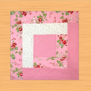 Puede incluir: Un bloque de edredón cuadrado con un diseño de cabaña de troncos. El bloque presenta un cuadrado rosa central, rodeado de tela floral blanca, luego tela rosa con un estampado floral de rosas rojas y hojas verdes. El borde exterior es rosa liso.