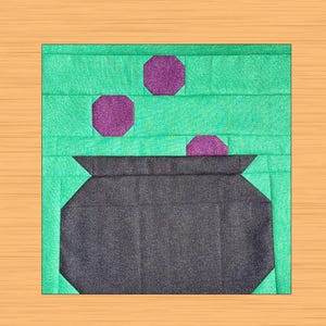 May include: A square quilt block featuring a dark gray cauldron with three purple octagons above it. The background is a light green color with horizontal lines. The block is set against a light brown wooden surface.