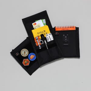Könnte beinhalten: Eine schwarze Reißverschlusstasche mit mehreren Taschen und Fächern. Die Tasche ist geöffnet und zeigt ein gelbes Notizbuch mit dem Schriftzug "Field Notes" auf dem Einband, ein Multitool, einen Stift, einen Kompass und ein paar Patches. Die Tasche hat auch ein kleines orangefarbenes Notizbuch mit dem Schriftzug "Defying" auf dem Einband.