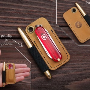 Pu&ograve; includere: Un organizzatore da tasca in pelle marrone con un coltellino svizzero rosso e una penna dorata. L'organizzatore ha un anello per un portachiavi e uno slot per una penna. Il testo "penslip is optional" &egrave; scritto sull'immagine.
