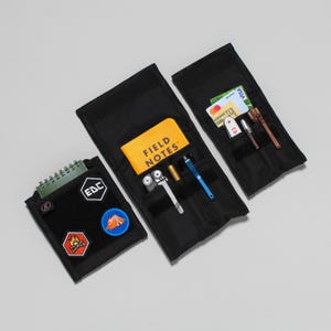 Pode incluir: Três bolsas pretas com zíper com diferentes conteúdos. A primeira bolsa tem um pequeno caderno com uma capa verde e três patches. A segunda bolsa tem um pequeno caderno amarelo com o texto "FIELD NOTES" e uma multiferramenta, uma caneta e uma pequena faca. A terceira bolsa tem um cartão de crédito, uma pequena faca e uma caneta.