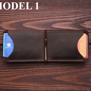 May include: A brown leather wallet with a money clip and two card slots. The wallet is open and has a blue card in the left slot and an orange card in the right slot. The wallet is on a wooden surface.