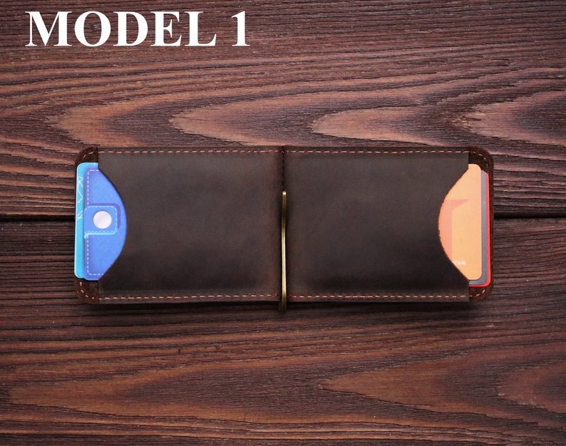 May include: A brown leather wallet with a money clip and two card slots. The wallet is open and has a blue card in the left slot and an orange card in the right slot. The wallet is on a wooden surface.