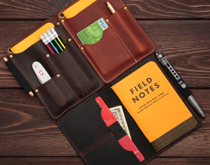 Puede incluir: Una cartera de cuero marr&oacute;n con m&uacute;ltiples bolsillos y ranuras. La cartera tiene una solapa de cuero negro con costuras rojas. La cartera est&aacute; abierta para revelar una libreta amarilla Field Notes, una tarjeta de cr&eacute;dito y un bol&iacute;grafo.