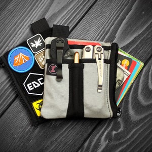 Op de afbeelding: Een grijze ritszak met zwarte bies en verschillende patches, waaronder een tent, een pootafdruk en "EDC". De zak is open en bevat een zwart vouwmes, een zilveren multitool, een pen en een stapel creditcards en contant geld.