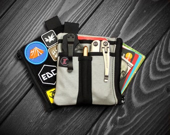 May include: A grey and black nylon pocket organiser featuring multiple exterior slots holding a black folding knife, a metallic pen, and a silver multi-tool. A separate black pouch with various colourful embroidered patches is attached behind it.