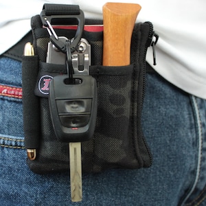 Puede incluir: Bolsa de cinturón utilitaria negra con un bolígrafo, multiherramienta, llave de coche y pequeña herramienta con mango de madera. La bolsa está sujeta a un mosquetón negro. La bolsa se lleva en la cintura, sujeta a unos vaqueros.