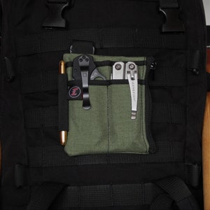 May include: An olive green fabric pocket organizer attached to a black tactical backpack. The organizer features two vertical slots holding a gold-toned pen and a black folding knife, plus a side compartment containing a silver multi-tool.