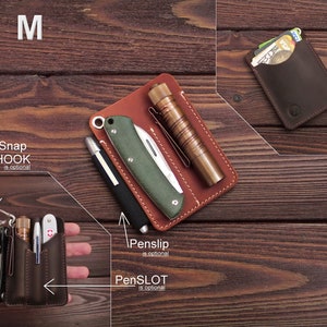 May include: A brown leather wallet with multiple pockets and slots for carrying cards, cash, and a pen. The wallet has a snap hook and a pen slip. The wallet is shown on a wooden surface.