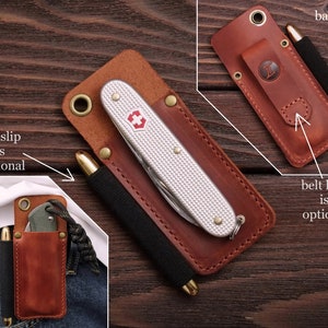 Pu&ograve; includere: Un portacoltelli e penne in pelle marrone con un coltellino tascabile argentato e una penna nera. Il portacoltelli ha un passante per cintura e una fessura per penne. Il testo "penslip is optional" e "belt loop is optional" &egrave; visibile nell'immagine.