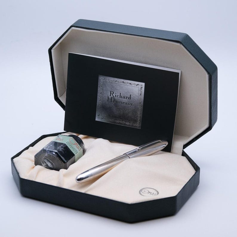 May include: A silver pen, ink bottle, and card are presented in a black octagonal box. The card reads "Richard Hennessy" in a silver font. The pen is sleek and metallic, and the ink bottle is black with a green label.