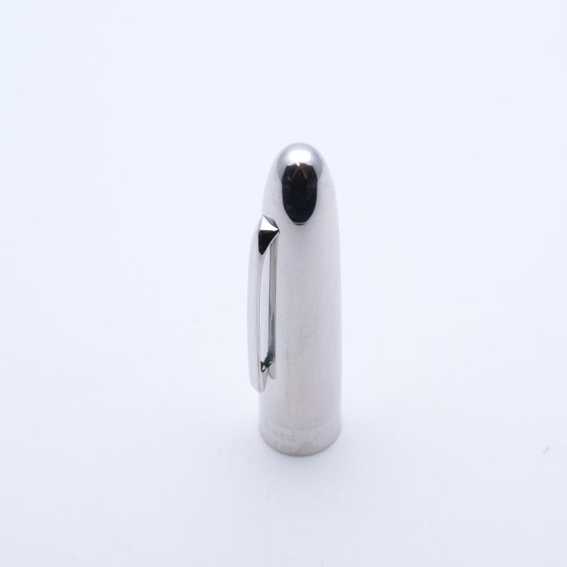 May include: A polished silver pen cap, featuring a rounded end and a small, rectangular clip. The cap has a cylindrical shape and reflects light, giving it a bright, metallic sheen. This pen cap is likely for a fountain pen or similar writing tool.
