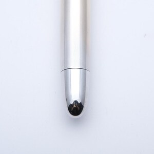 May include: Close-up of a silver pen with a rounded tip. The pen's body has vertical lines, and the tip reflects the surrounding environment. The pen is set against a white background.