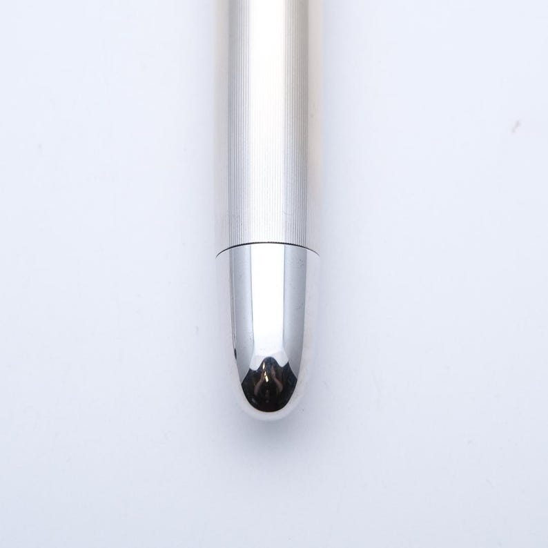 May include: Close-up of a silver pen with a rounded tip. The pen's body has vertical lines, and the tip reflects the surrounding environment. The pen is set against a white background.