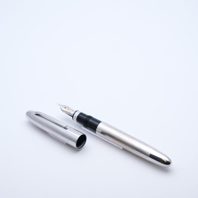 May include: A silver fountain pen with a black grip and a silver cap. The cap is lying next to the pen. The pen has a silver body and a silver nib. The pen is on a white background.