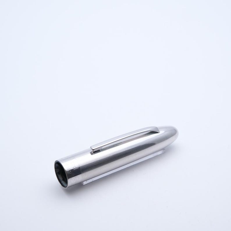 May include: A silver pen with a rounded end and a clip. The pen has a smooth, metallic finish and a cylindrical body with fine vertical lines. The pen is resting on a white surface.