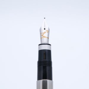 May include: Close-up of a fountain pen nib with gold detailing. The pen has a silver and black barrel. The nib is silver with gold accents. The pen is designed for writing and calligraphy.